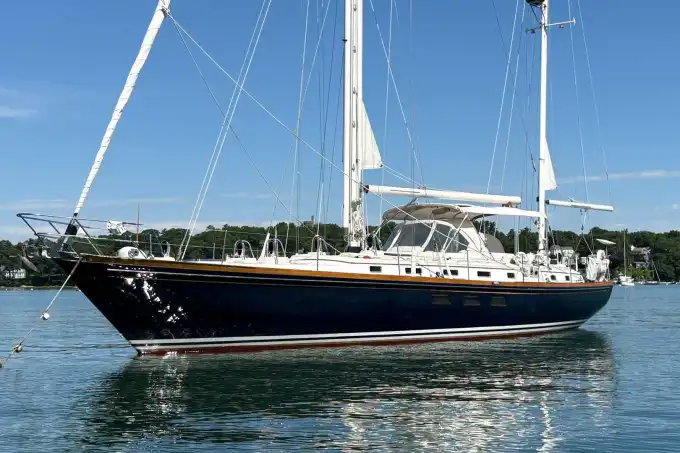 A large, elegant sailboat with white sails and a dark blue hull floats on calm water near a shoreline with green trees under a clear blue sky.
