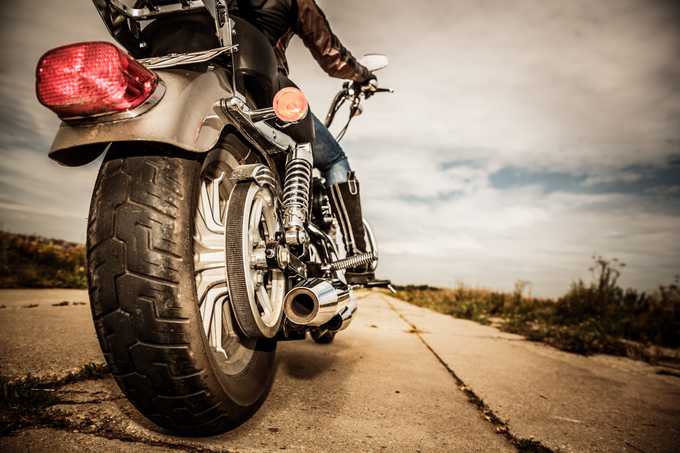 Low-angle view of a person riding a motorcycle on a rural road, focusing on the rear wheel, exhaust, and taillight, with cloudy sky and open landscape in the background.