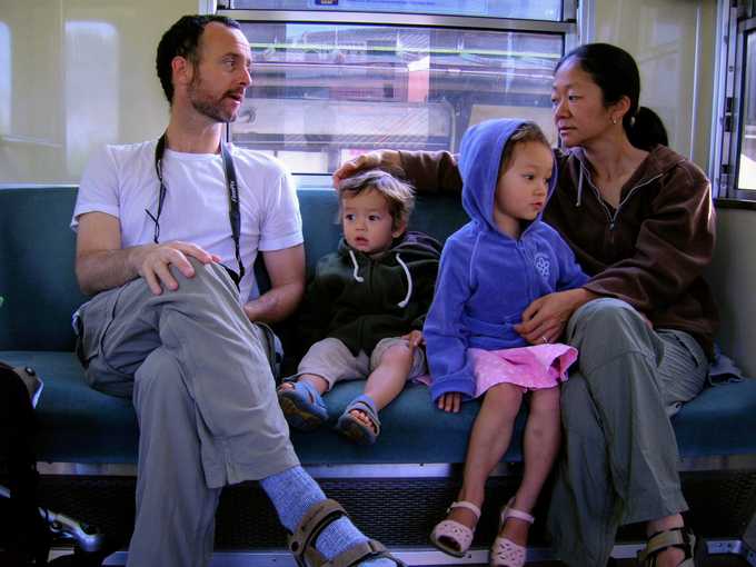 Four people, two adults and two children, sit together on a bench inside a train. The adults look at each other while the children sit quietly in between them, dressed in casual clothing.