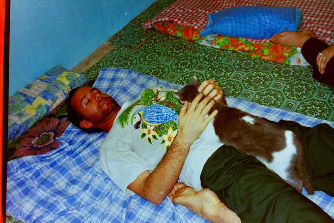 A man lies on a patterned blanket indoors, petting a gray and white cat resting on his chest. Another persons feet are visible nearby on a separate blanket.
